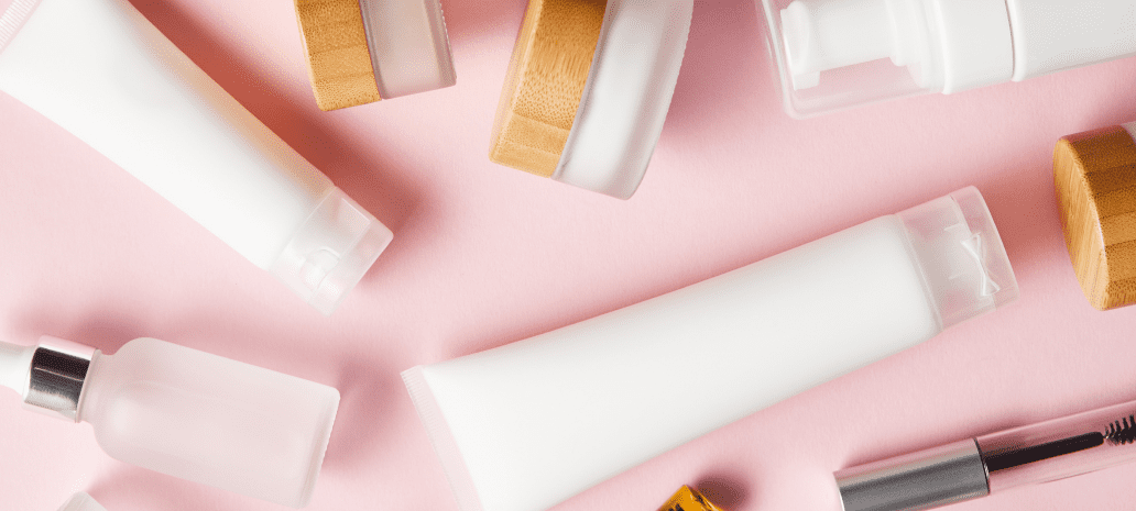 Skincare and cosmetic products in minimalist white packaging with wooden accents, arranged on a soft pink background.