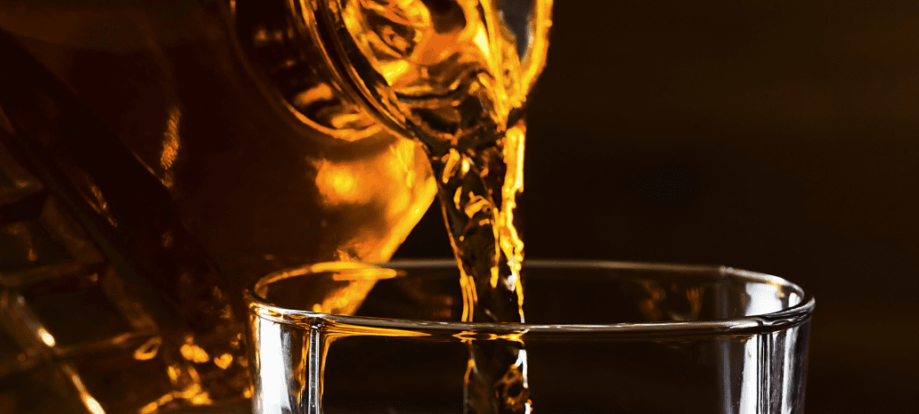 A glass filled with ice as whiskey is poured from a decanter, captured in warm lighting on a wooden surface.